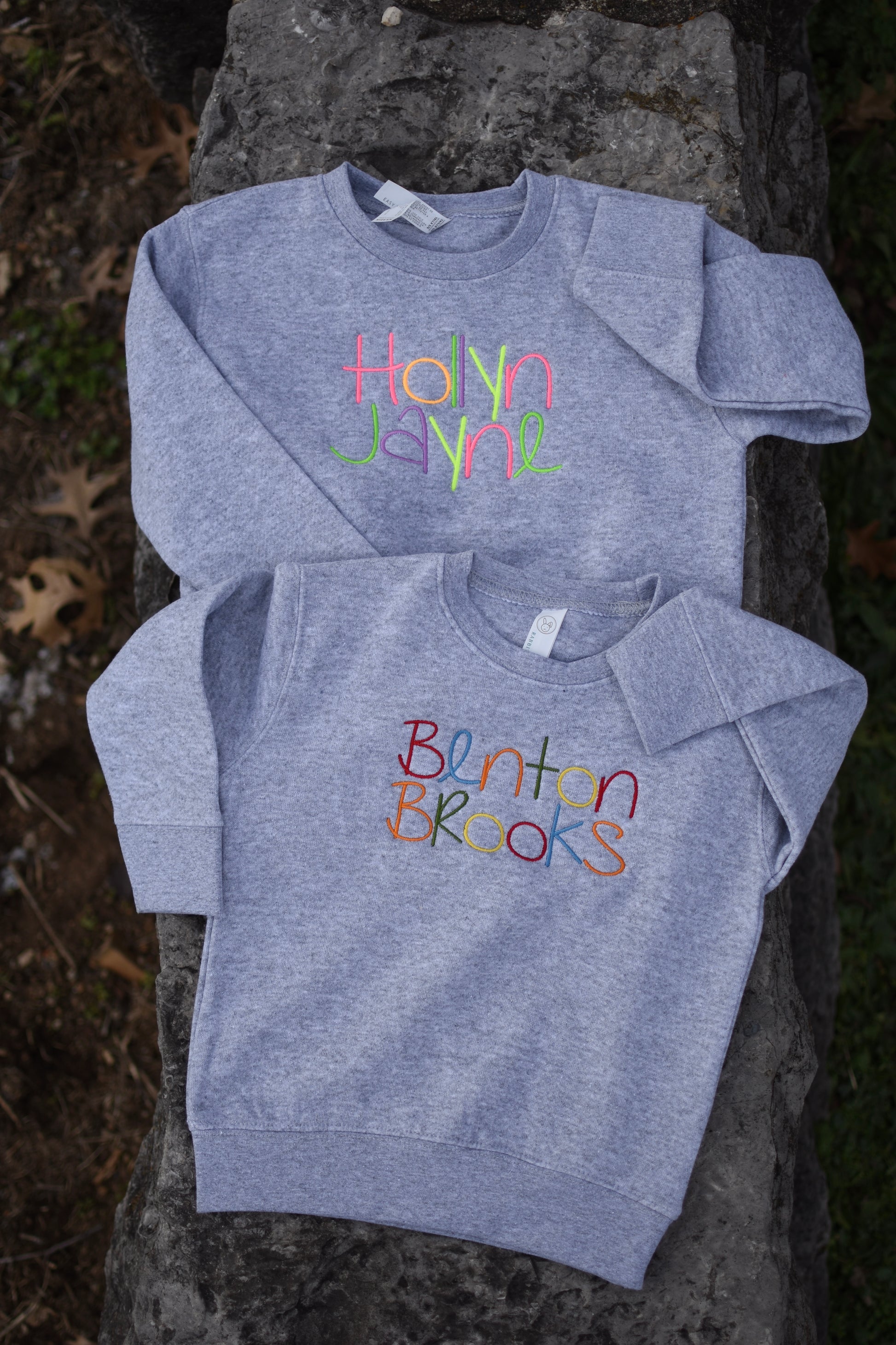 Two gray sweatshirts with colorful text on the front, one with the name 'Hayden' and the other with the name 'Benton'. Both are placed on a stone surface outdoors.